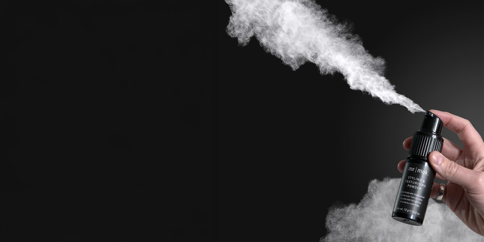 Hand pushing down on the mr muk Styling & Texturising Powder pump spraying white powder on a black background